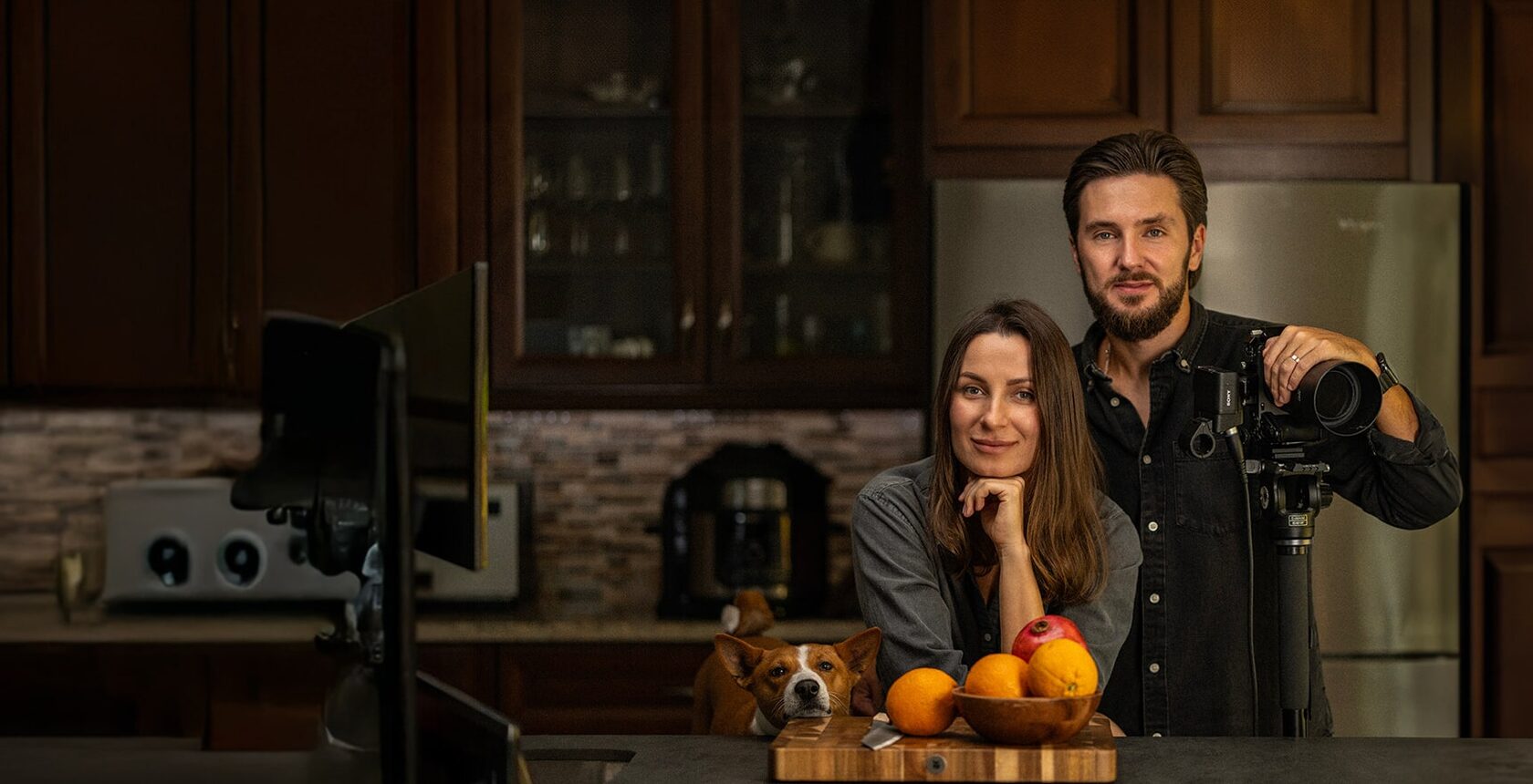 Max and Victoria in their kitchen: he holds a camera on a tripod, a board of fruit in front of them, their dog beside